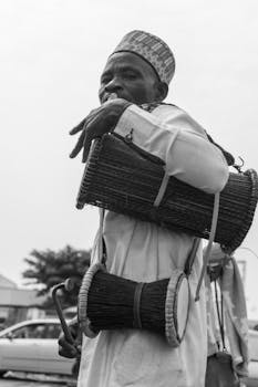 Black and white photo of a man in traditional attire playing drums outdoors in Abuja, Nigeria.