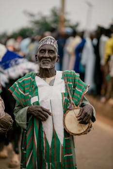 Elderly African man playing drum at cultural event outdoors.
