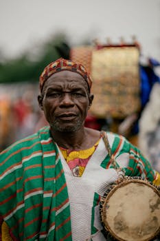 Elderly African man in colorful traditional attire playing a drum at an outdoor cultural event.